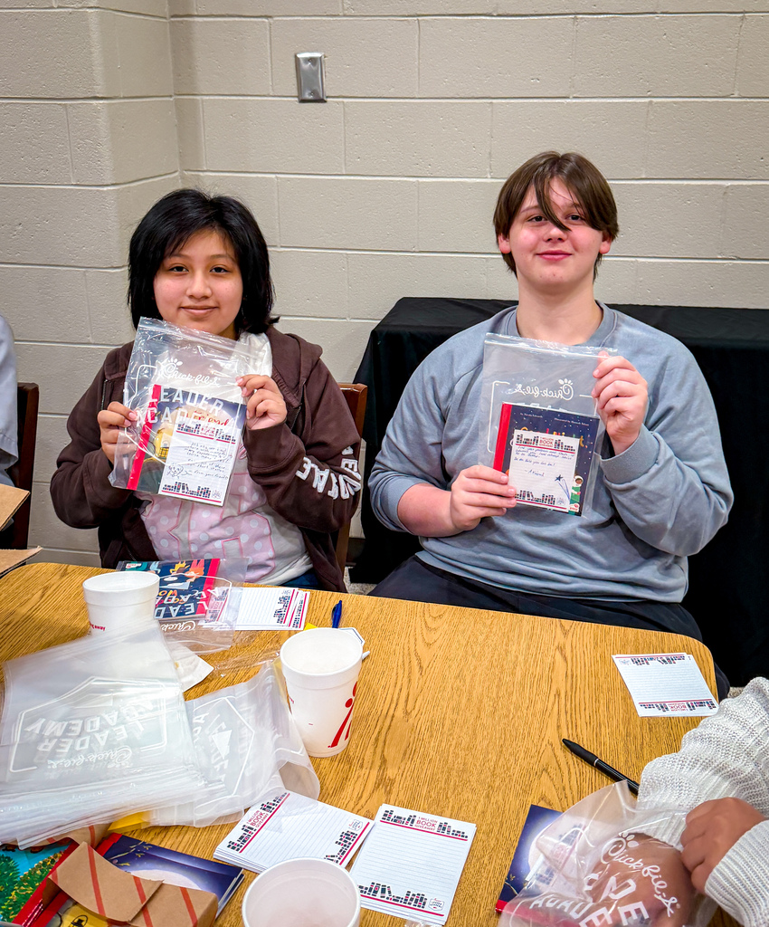 Two individuals sitting at a table hold plastic bags with printed materials inside. One wears a brown shirt, the other a blue one.