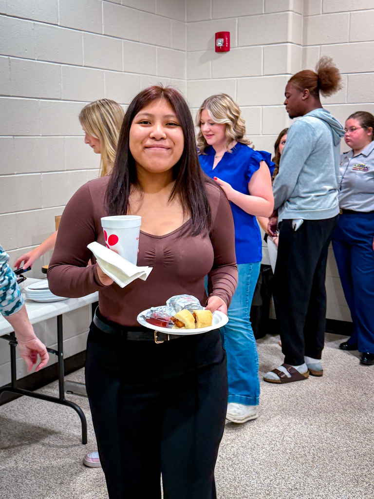 A woman holding a paper cup and a plate of food stands in a room with other people.
