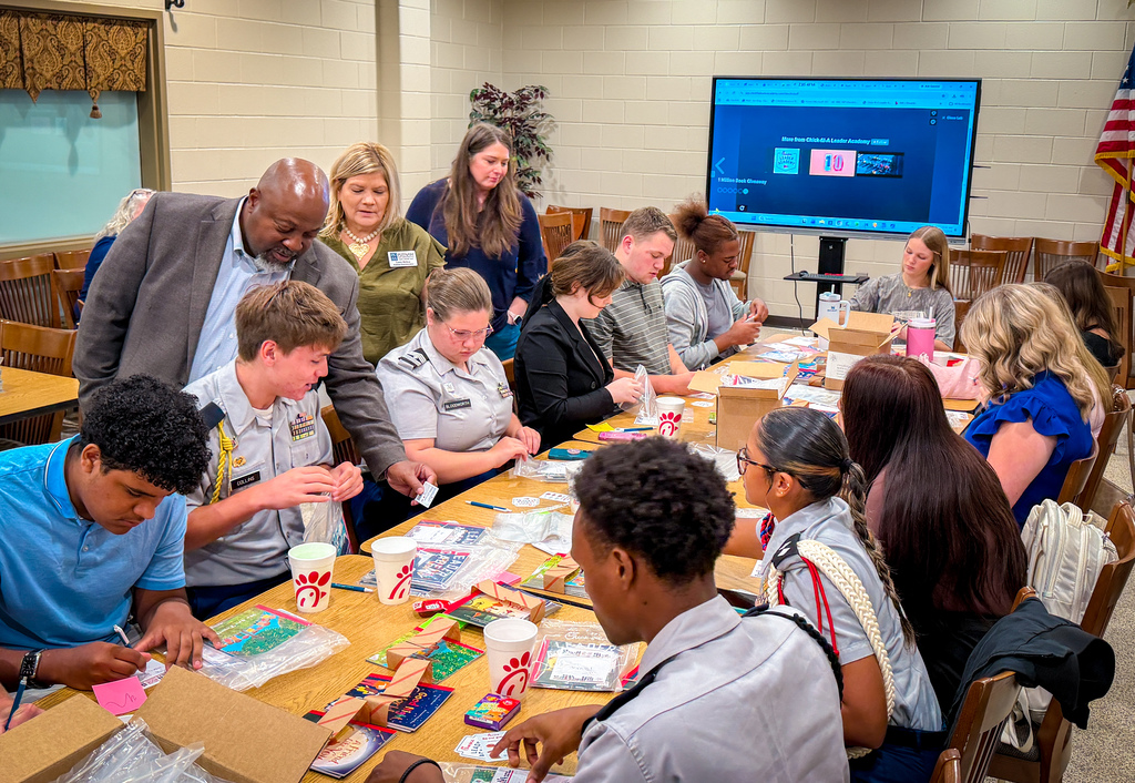 People at a table with craft supplies, looking at a screen, with one person talking.