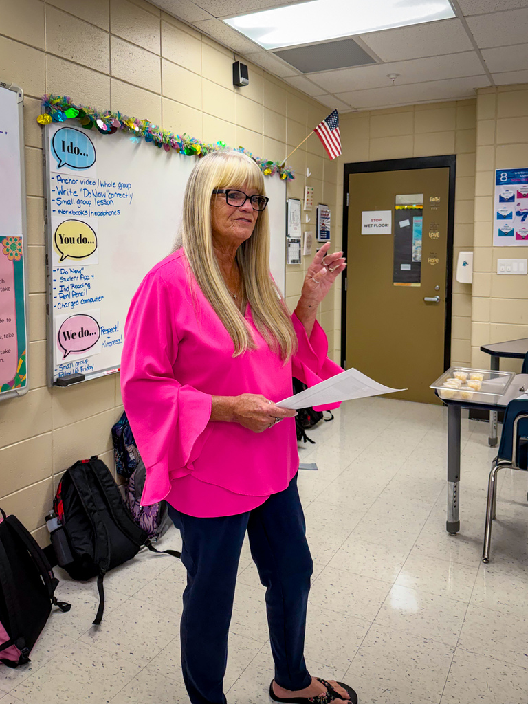 A woman with blonde hair stands in a classroom holding paper and speaking. A whiteboard and flag adorn the background.
