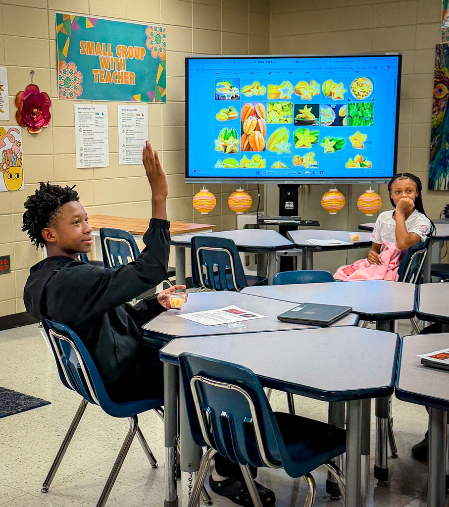 Two students seated in a classroom. One raises their hand. A screen displays a food-themed presentation.