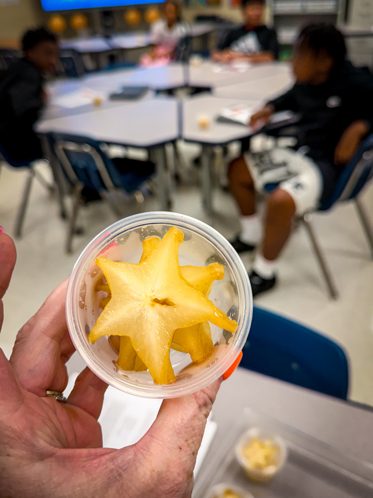 Hand holding a container of star fruit in a classroom. Other students are seated at tables.
