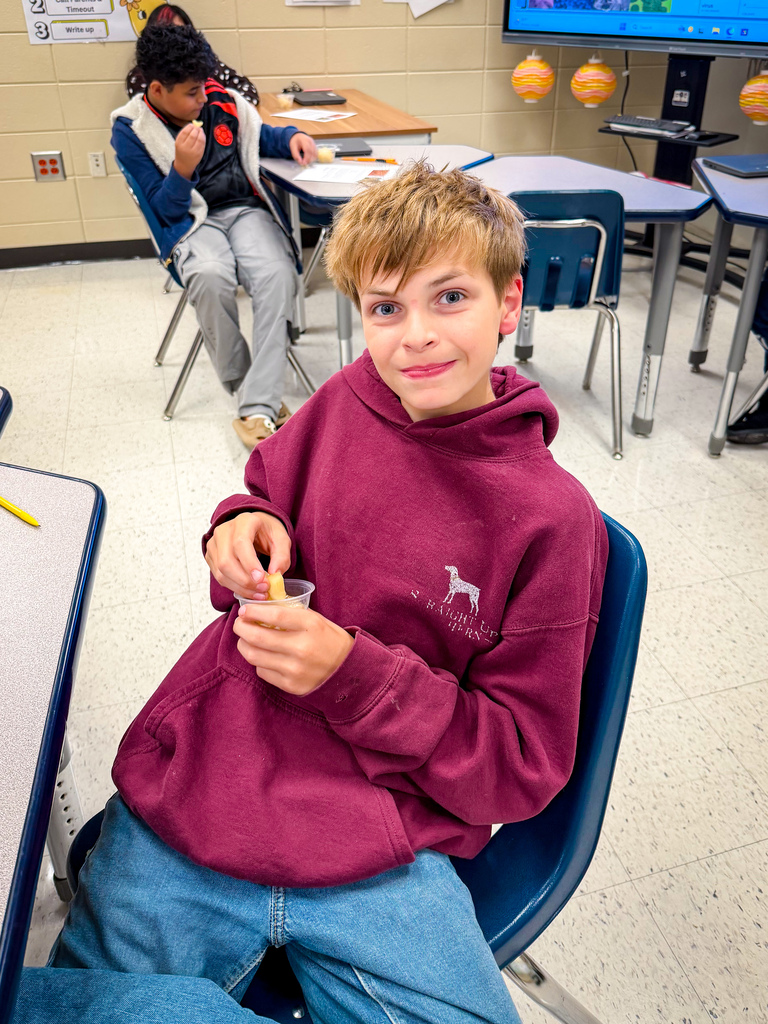 A boy in a maroon sweatshirt sits in a chair in a classroom, holding a small container.