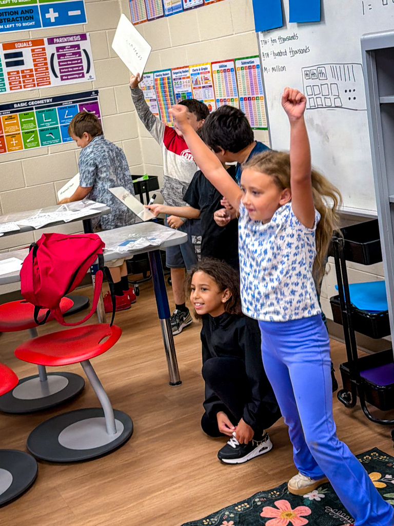 A classroom with kids standing and sitting. A girl in a floral shirt raises her arms while others hold papers.