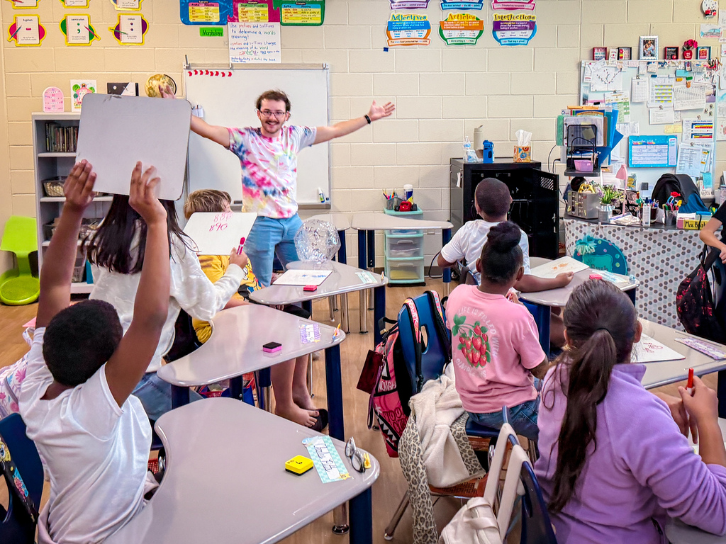 A classroom with children seated at desks. A teacher stands with raised arms. Desks have books, papers, and supplies.