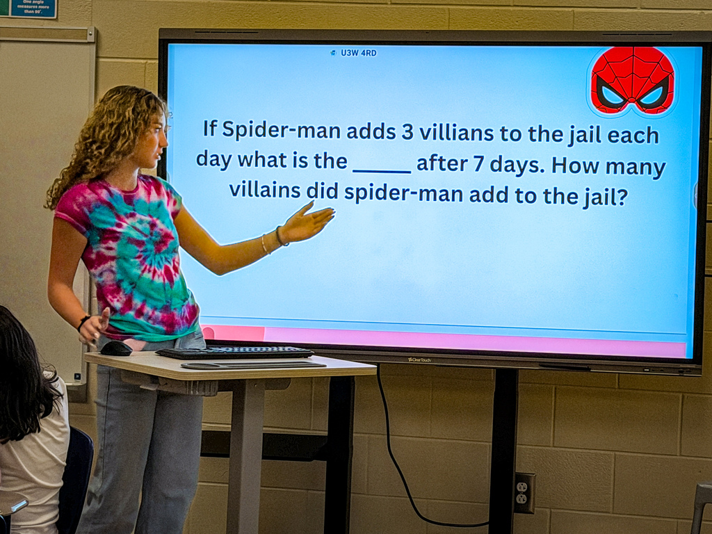 A woman stands at a podium in a classroom, pointing to a large screen showing a math problem.