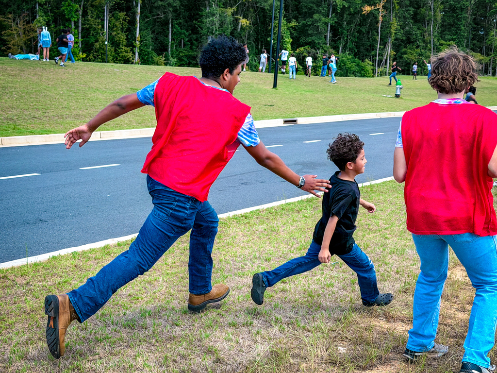 A child holds hands with two adults in red vests as they run across grass by a road.