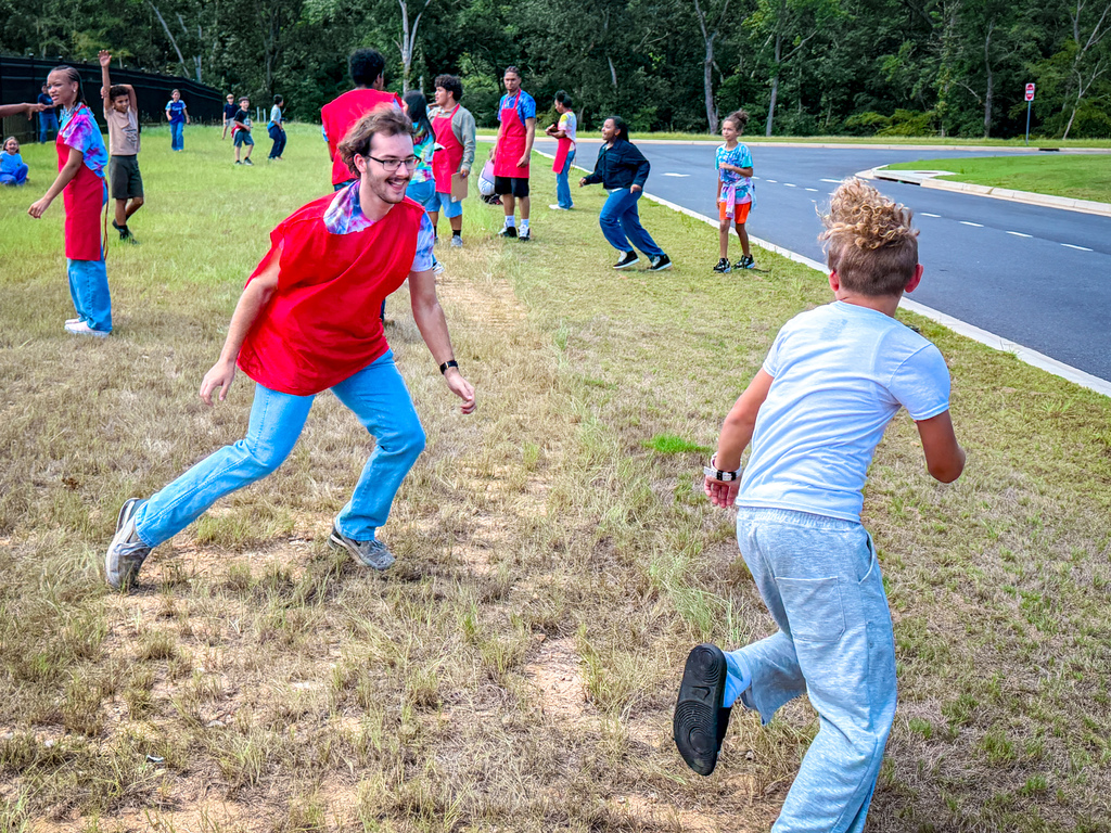 Multiple children run on a grassy area. A man in red wears glasses. Another child in white runs ahead.