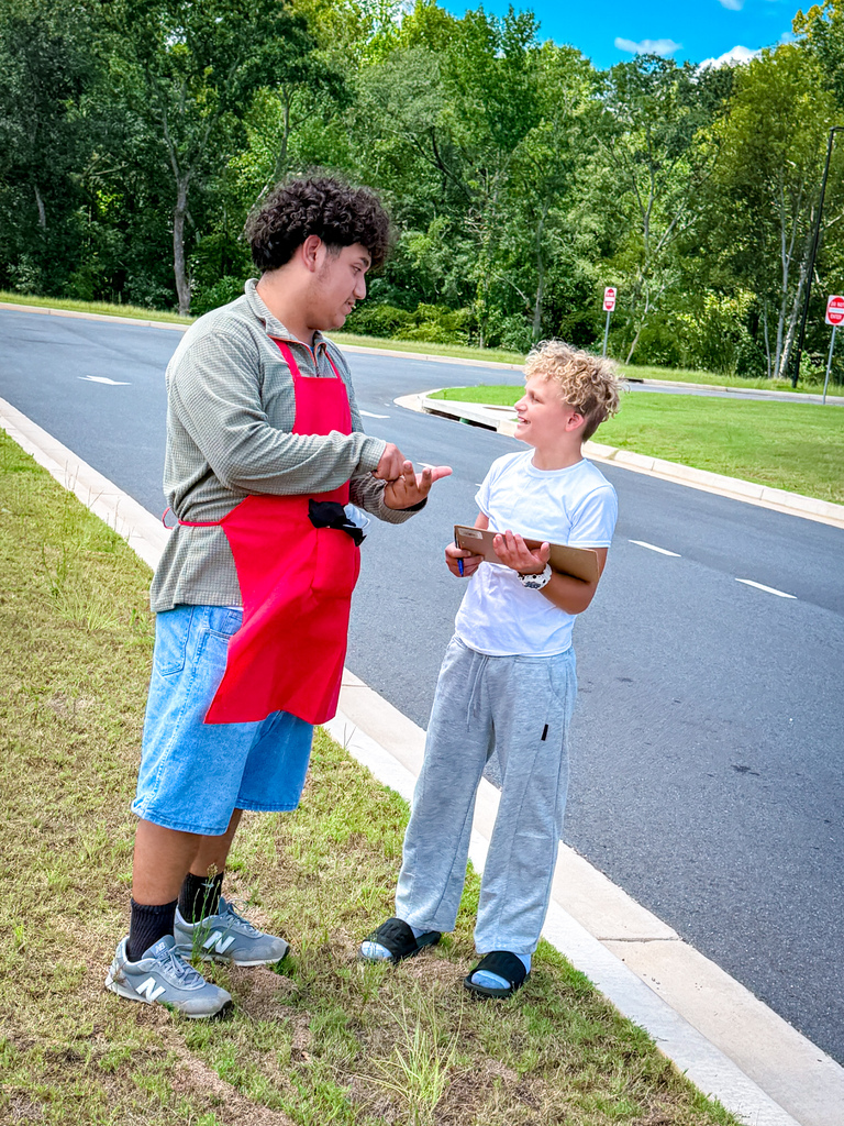 Two individuals standing on grass beside a road. One wears an apron; the other holds a chocolate bar.