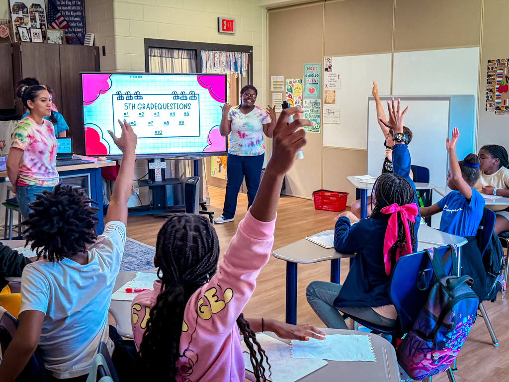 A classroom with children raising hands. A teacher stands before a large screen and gestures. Brightly colored decor on walls.