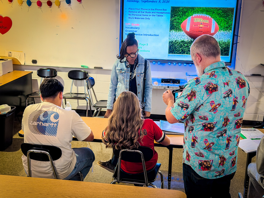 People in a classroom. A man and woman work with a young girl. A large screen in background displays a football.