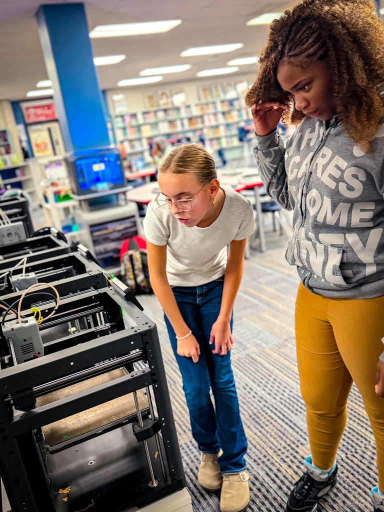 Two women examine a 3D printer in a library. A woman in white stands next to the printer.