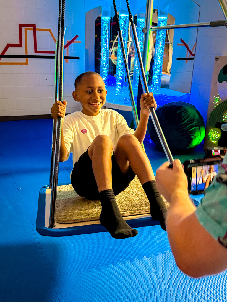 A smiling child in a white shirt and black shorts swings on a swing with colorful lights in a colorful room.