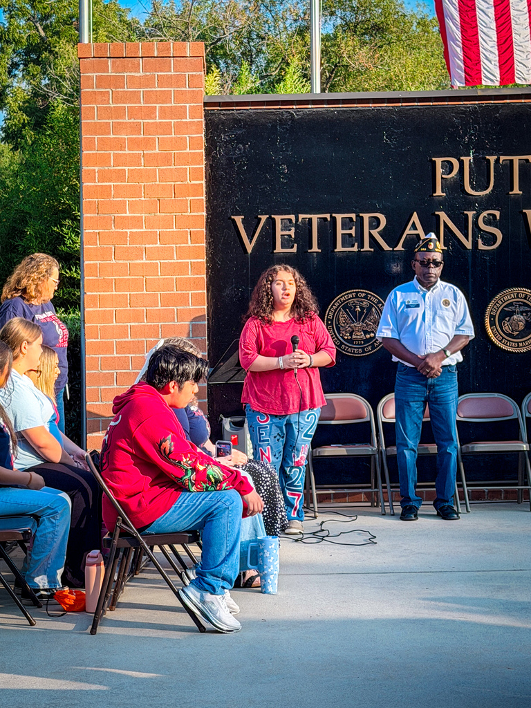 People seated at chairs, two standing in front of a podium, one holding a microphone. A sign reading "Put Veterans" is behind them.