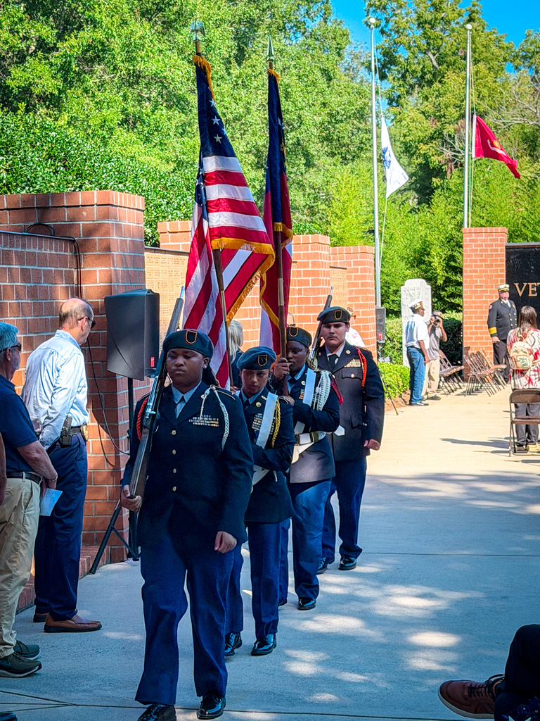 PCHS JROTC Cadets carrying flags walk past a brick wall with a speaker to the left. People stand along the sides.