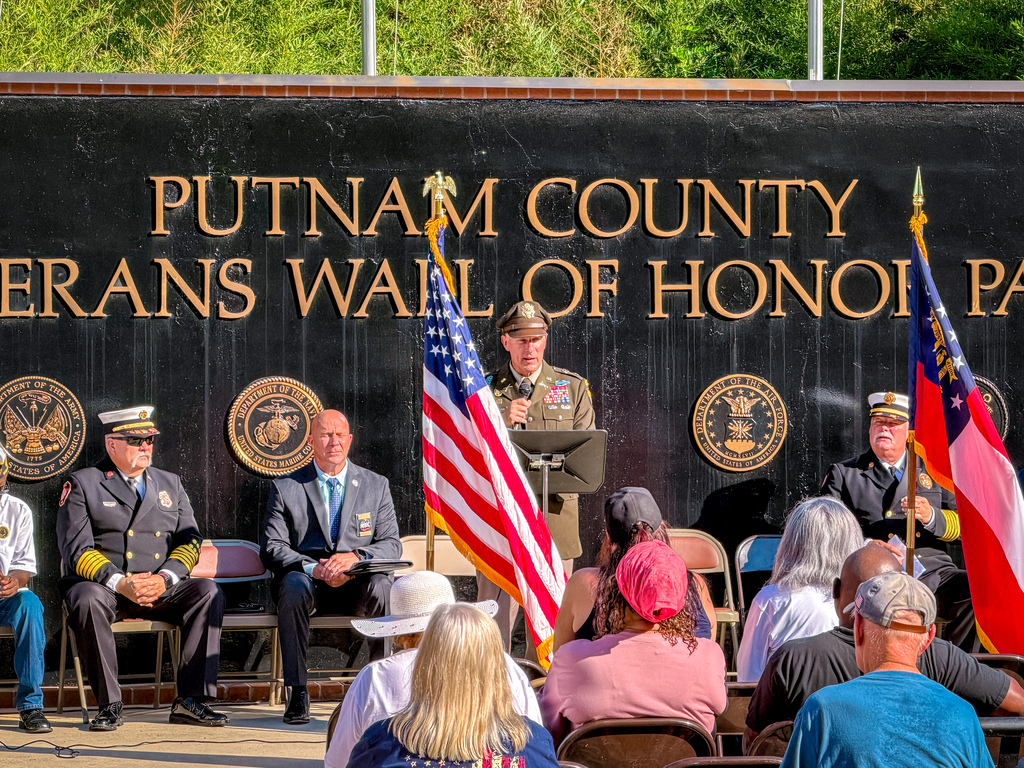 A group of people gathered at an outdoor event. In front is a podium with an American flag. Behind it is a wall with text.