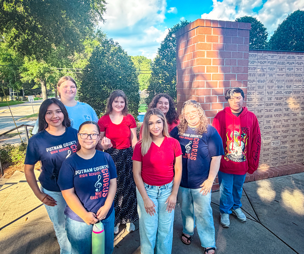 PCHS Chorus students wearing matching blue shirts stand in front of a red brick wall, surrounded by trees.