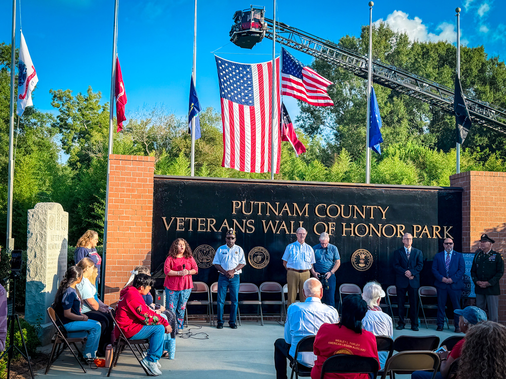 A group gathers in front of a brick wall. The wall displays the words "Putnam County Veterans Wall of Honor Park".
