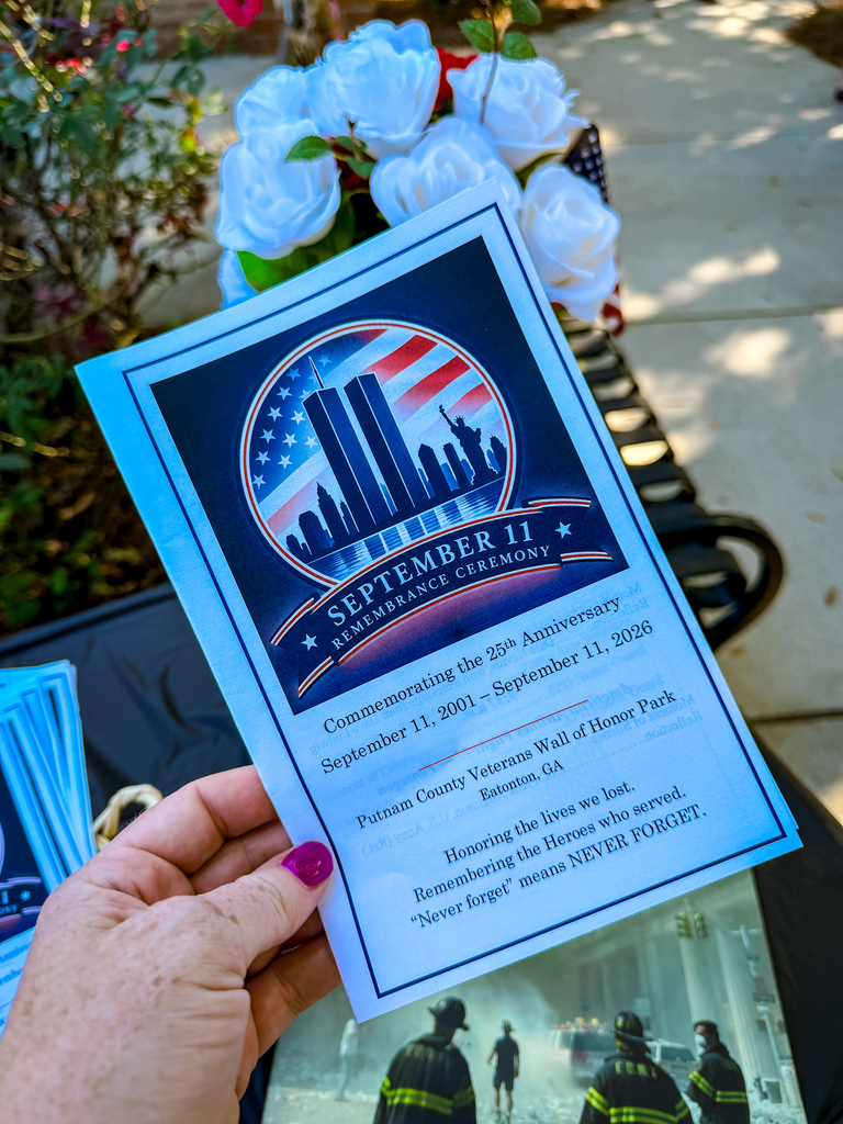 A hand holds a blue flyer titled "September 11 Remembrance Ceremony." The flyer features a cityscape and American flag.