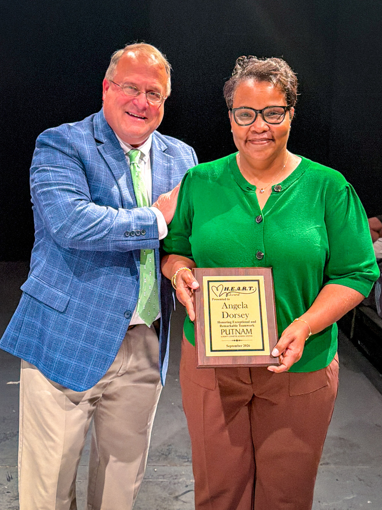 A man and a woman stand together, smiling. The man is in a blue suit and green tie, and the woman holds an award plaque.