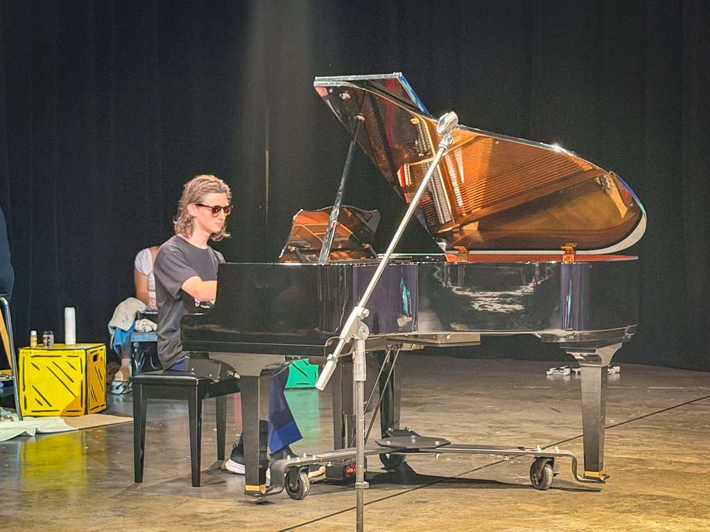 A person wearing glasses sits at a grand piano with the lid open, a microphone nearby. A person sits behind them.