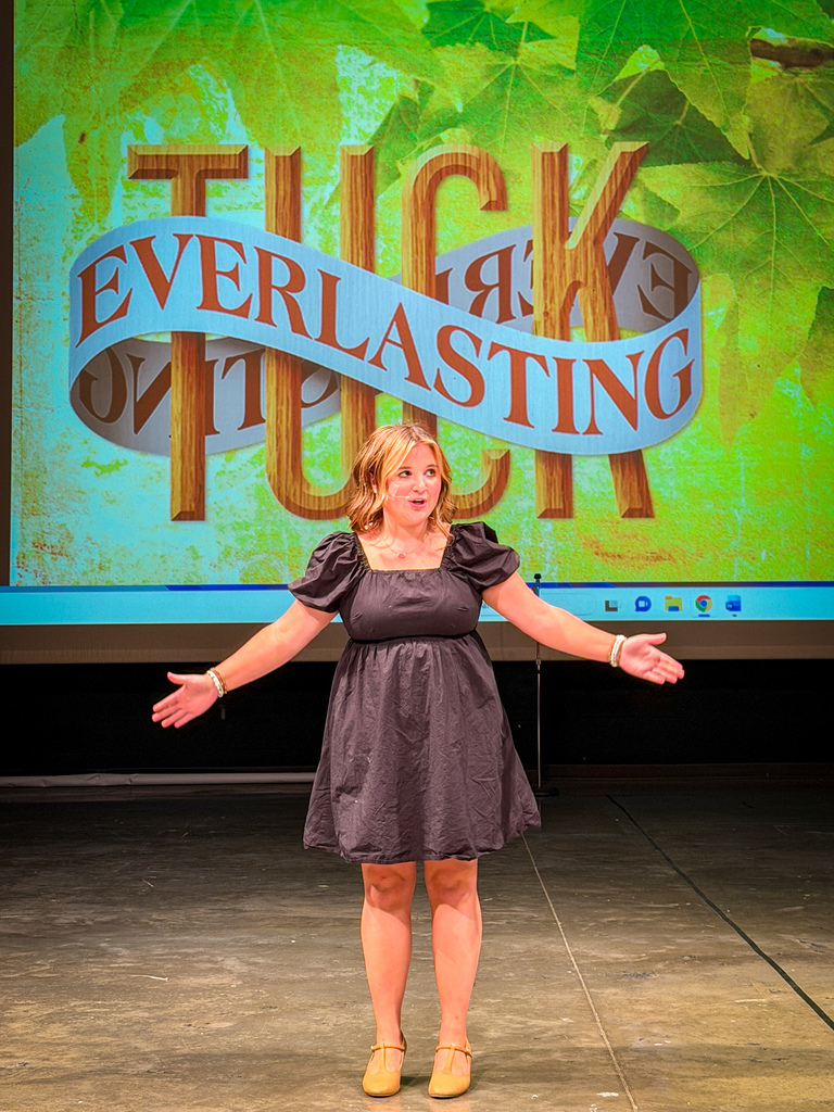 Woman in a dress stands on stage with arms extended. Green screen with text reads "TUCK EVERLASTING" in large letters.