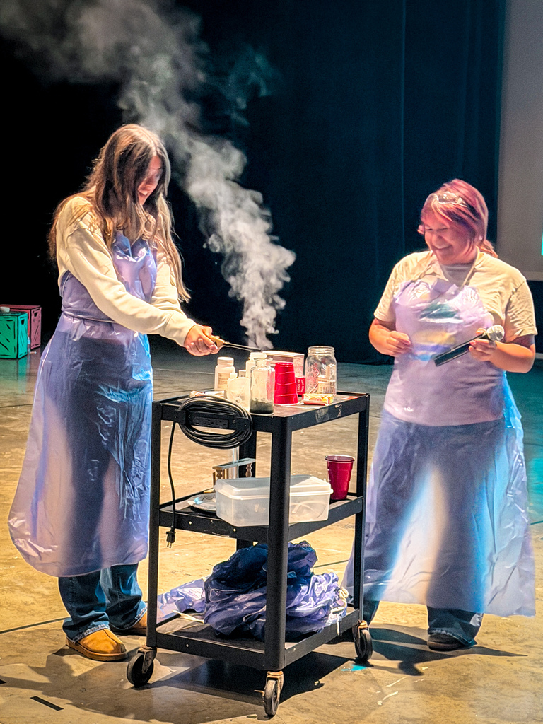 Two women in lab coats and plastic aprons standing by a cart with bottles and containers, one woman holds a lighter.