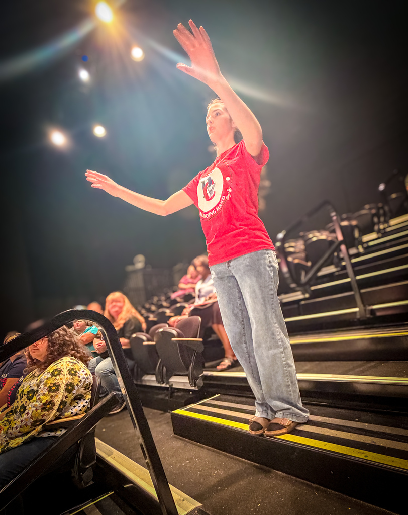 A person stands on stage with arms raised, wearing a red shirt and jeans. Below, an audience sits on stairs.