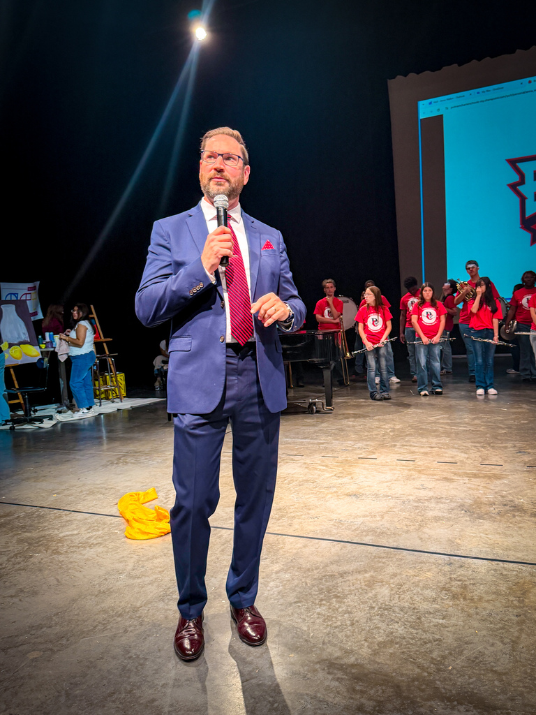 A man in a blue suit speaks into a microphone on a stage with a band in red shirts.