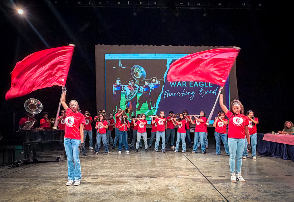 Two individuals in red tops and blue jeans hold up red flags on a stage. Behind them, a band performs, and a screen shows a marching band.
