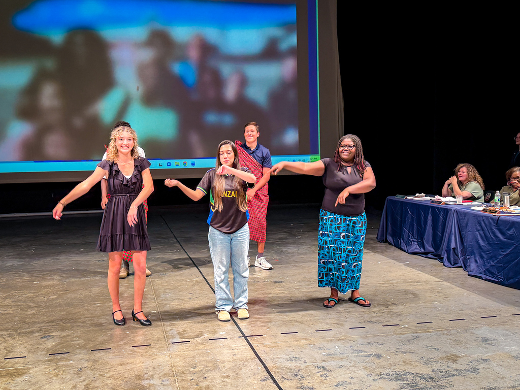 Five people on a stage with a video screen. One person wears a red garment, another has a blue skirt.