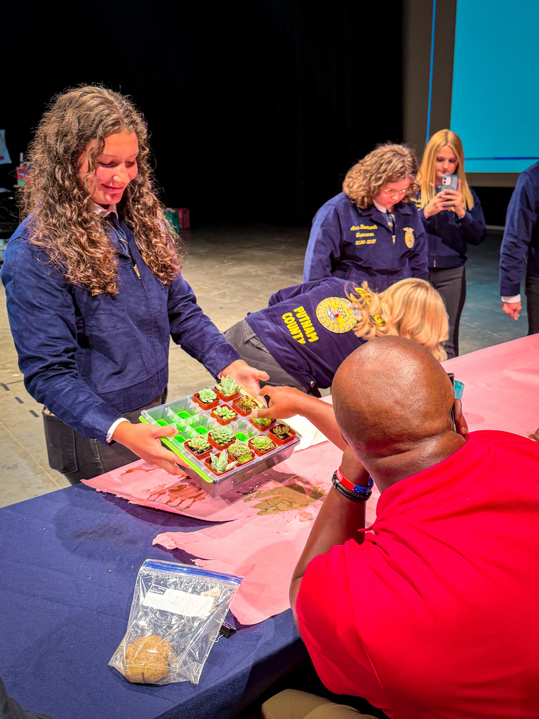 Four women in uniforms prepare to give a man a tray of cupcakes. The man sits at a table.