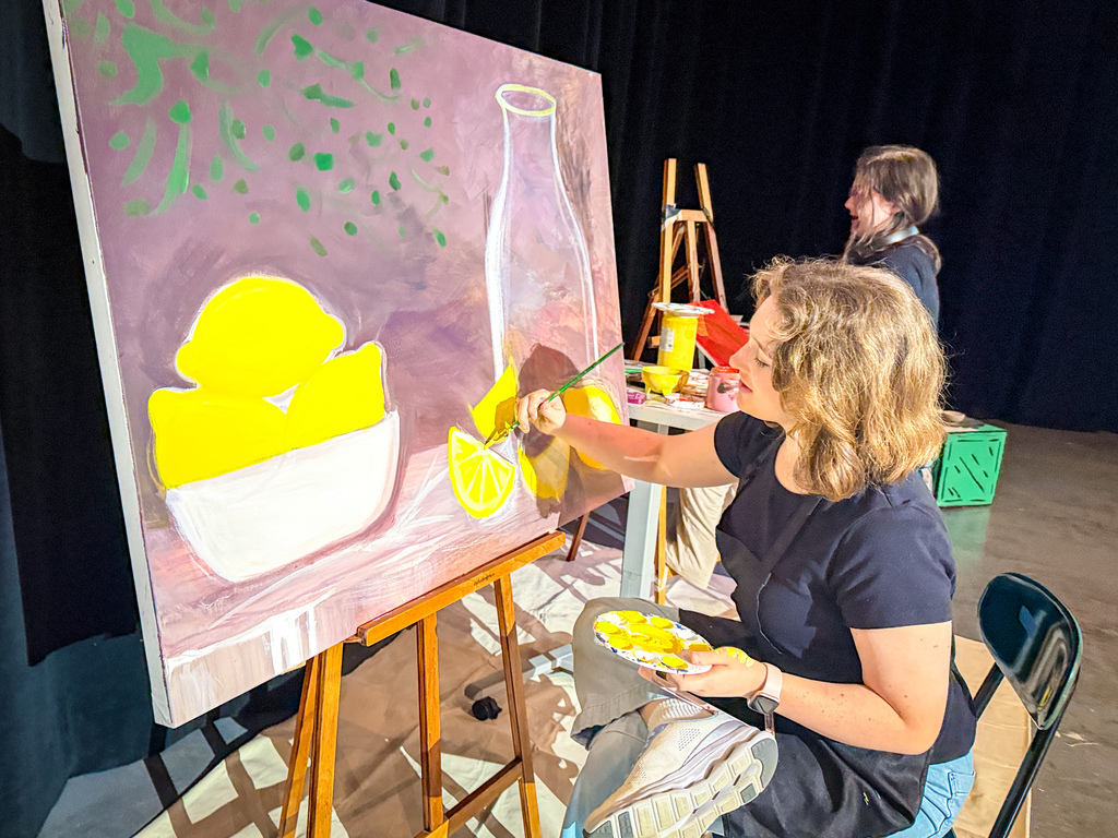 A woman painting a bowl of lemons on a large canvas on an easel, seated in front of a black curtain.