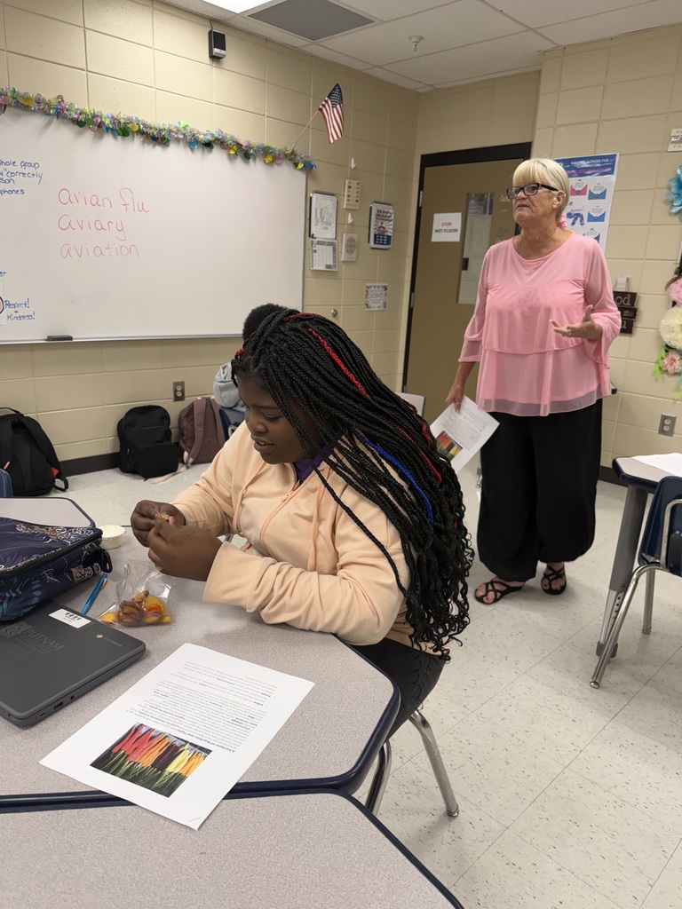 A woman with braided hair sits at a desk in a classroom with another woman standing nearby.