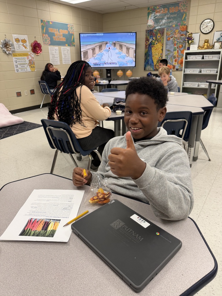 A student gives a thumbs up in a classroom, seated at a desk with a laptop and a pencil.