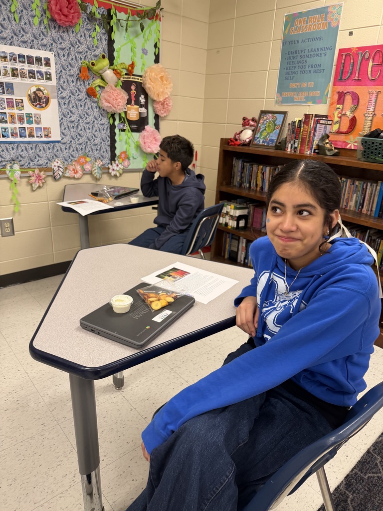 A classroom with a student in a blue hoodie seated at a table with books and a tray, another student sitting in the background.