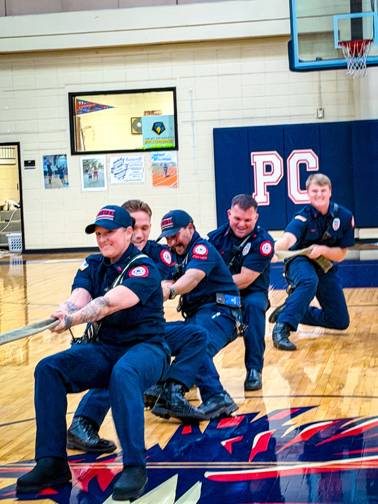 Six firemen in blue uniforms pull on a rope in a gymnasium. A blue banner with white letters reads "PC".