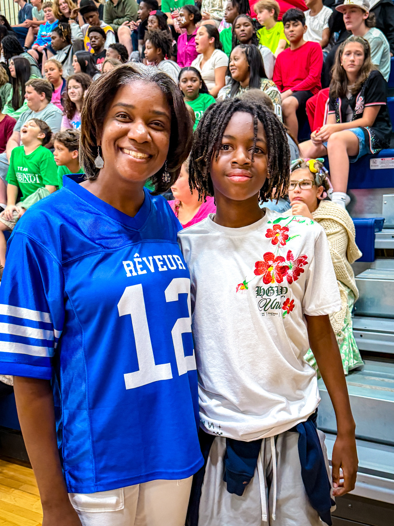 Woman and girl standing, smiling at camera. Audience seated on bleachers behind. Brightly colored shirts with numbers and designs.