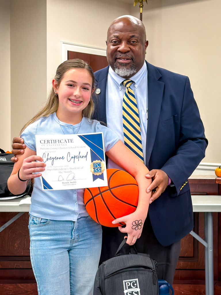 A girl in a blue shirt holds a certificate and a basketball. A man in a suit stands behind her.