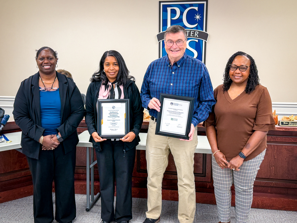 Five adults stand with framed certificates. A sign with blue and white letters reads "PCS" in the background.