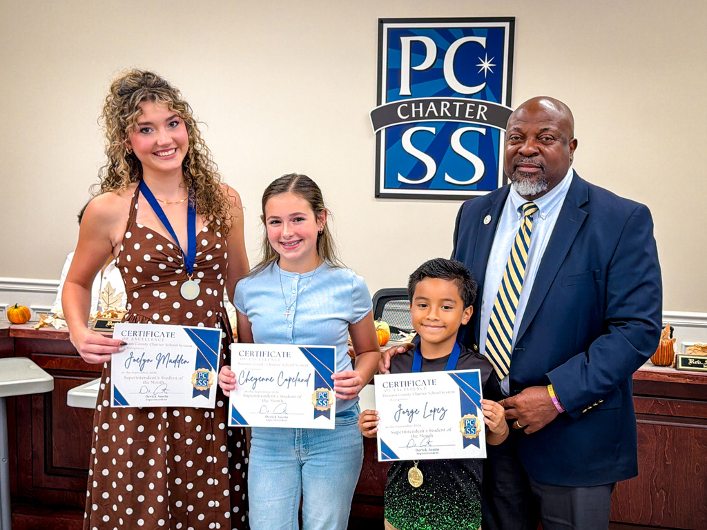 Three children and a man hold certificates in front of a sign reading "PC Charter SS".