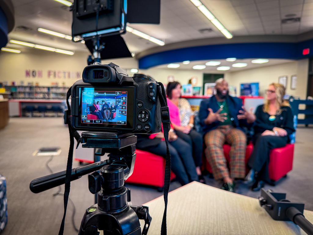 A camera on a tripod records a discussion. Three individuals sit on a red couch. A table holds a microphone.