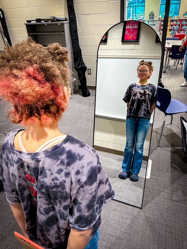 A young person with curly, pink-dyed hair and a tie-dye shirt views themselves in a mirror.