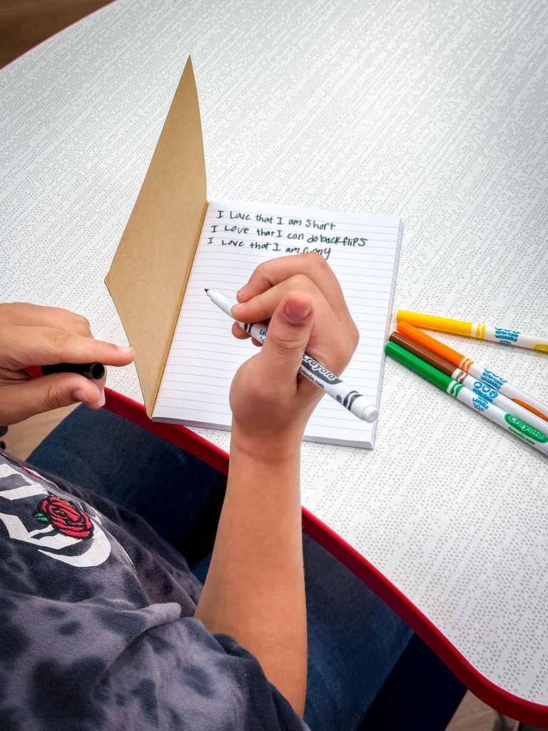 A person at a table with a pen, notebook, and markers. Writing on the open notebook.