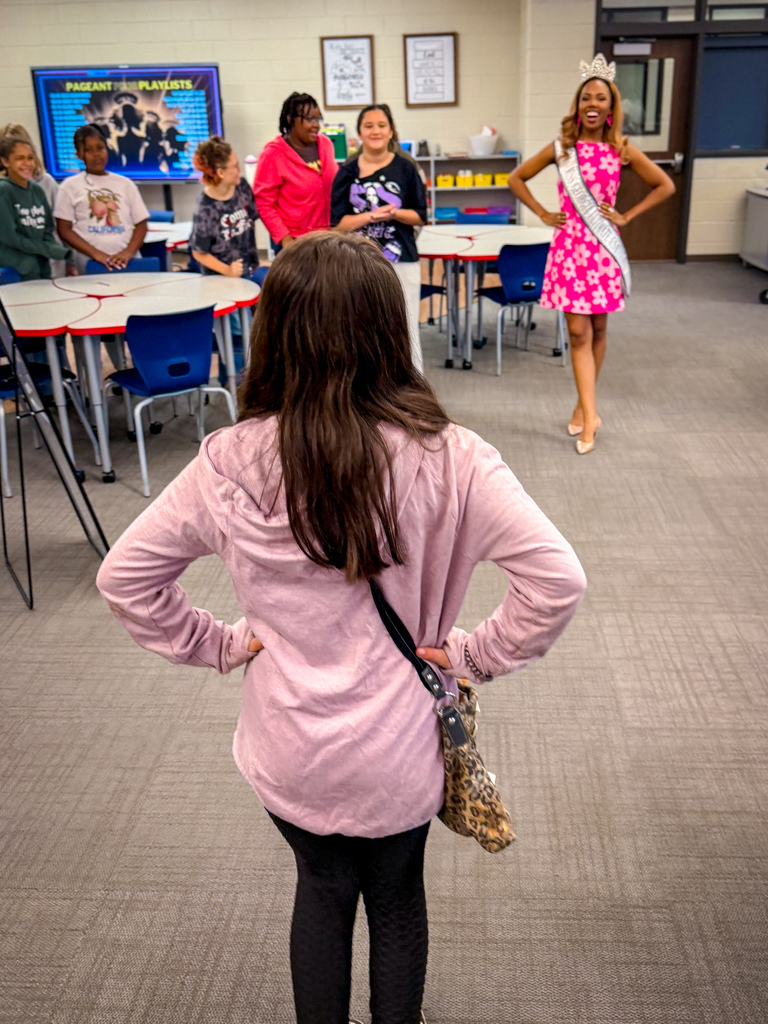 A classroom with tables, chairs, and people. A girl in a pink jacket faces away while others stand nearby.