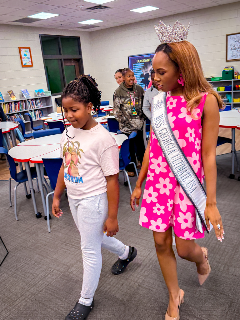 Two young girls walk inside a classroom. One wears a tiara and sash, while the other has a T-shirt.