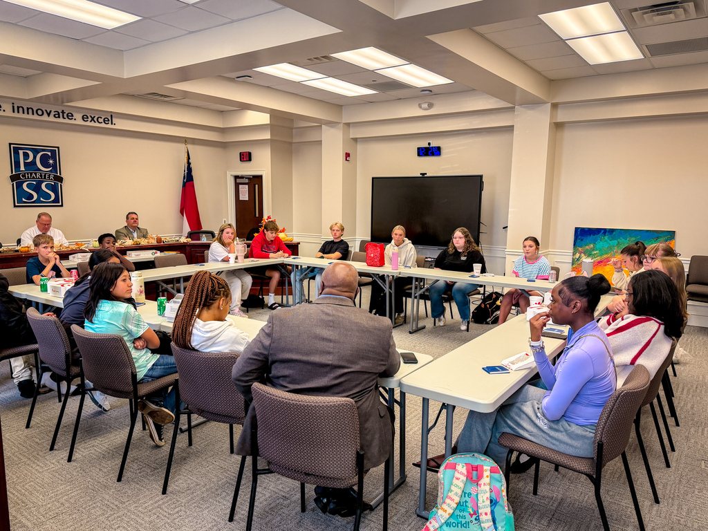 A conference room with people seated around tables. A flag is displayed on the wall. A monitor is on the wall.