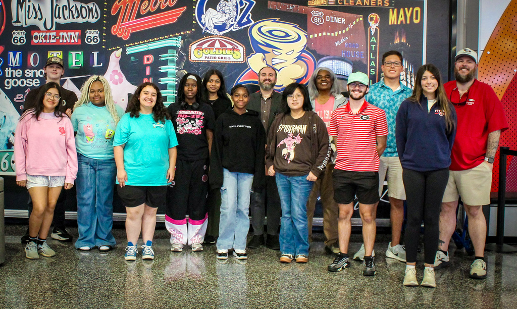 A group of people posing for a photo with a colorful, sign-adorned wall in the background.