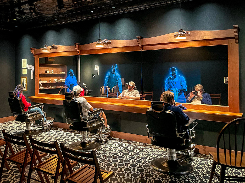 A room with black walls and patterned floor has chairs around a bar. Five people sit in chairs facing a mirror.
