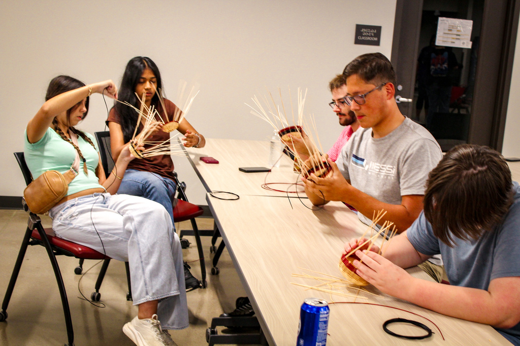 Five people seated around a table, engaged in crafting with straw. A woman in white sneakers holds a brown bag.